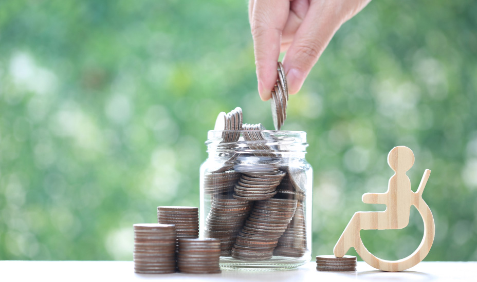 Hand places coins into a jar to save money next to wooden figurine of wheelchair